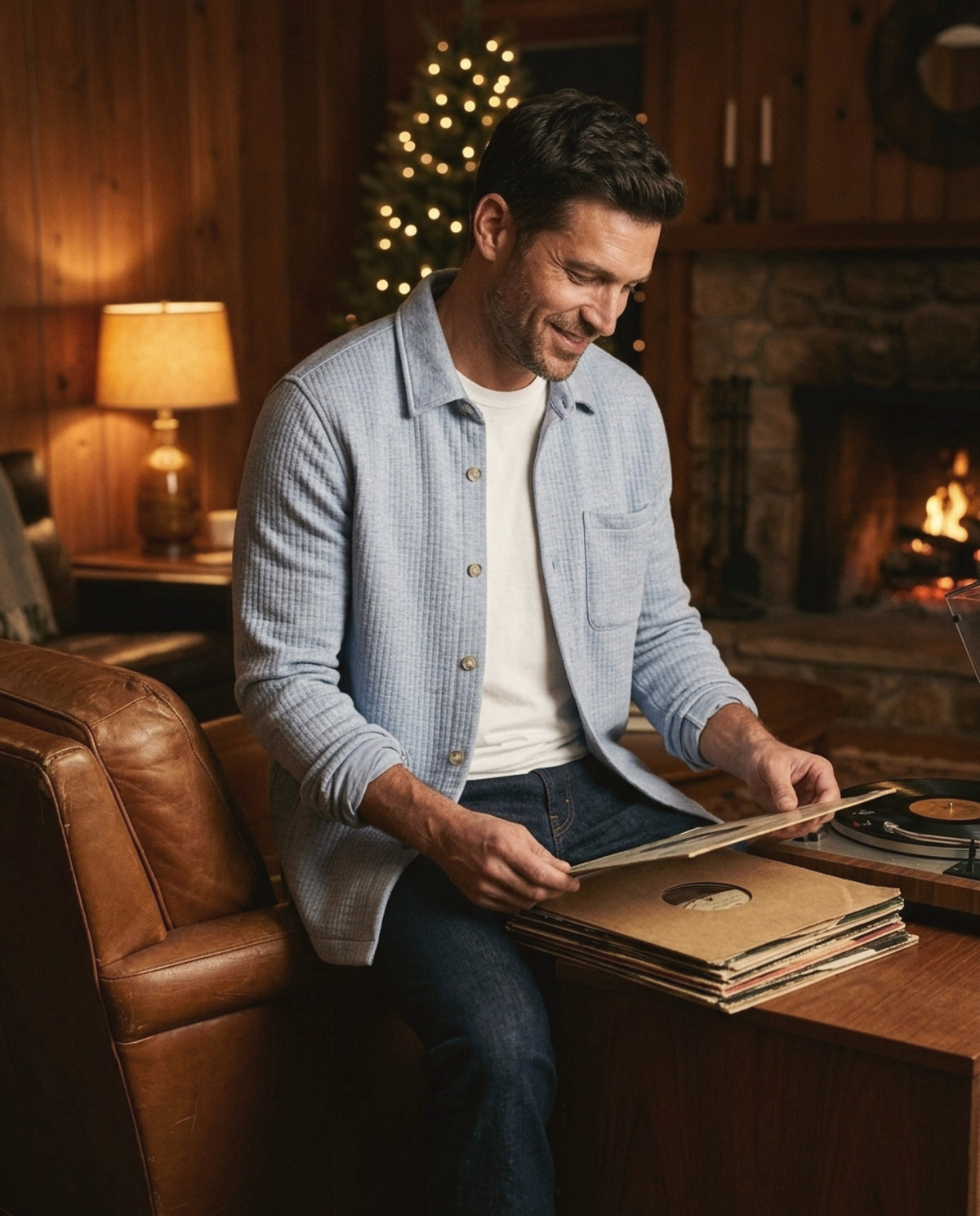 Man in a pale blue quilted overshirt looking through vinyl records by a fireplace