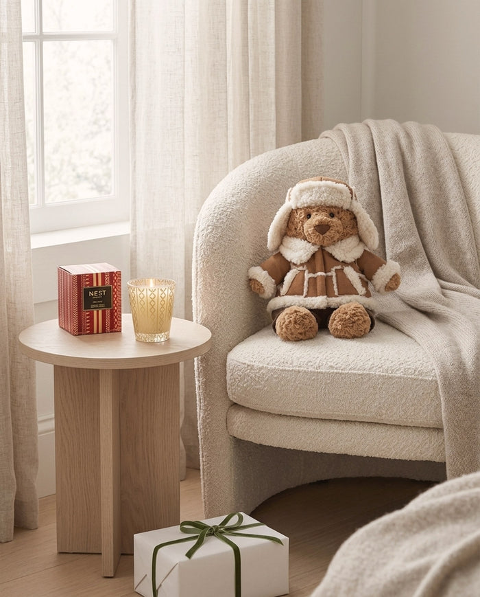 Holiday gifts under $100: Jellycat Bartholomew Bear Alpine on an armchair beside a lit Nest Holiday Classic candle