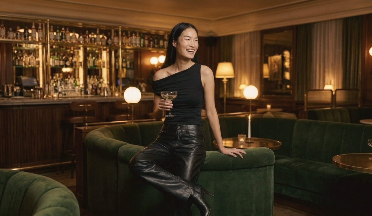 Woman in a black one-shoulder top and black leather pants laughing with a glass of champagne in a candlelit lounge
